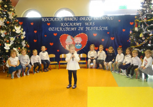 Amelka stoi na środku holu, trzyma mikrofon i mówi wiersz. Dzieci siedzą w półkolu.