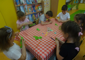 Sześcioro dzieci siedzi przy stoliku, owijają zieloną bibułą patyki