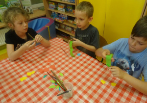 Troje dzieci siedzi przy stoliku i obijają patyczek zieloną bibułą