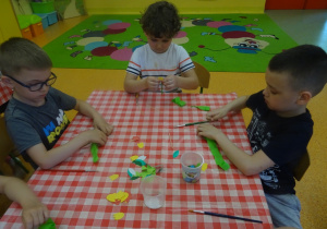Troje dzieci siedzi przy stoliku i obijają patyczek zieloną bibułą