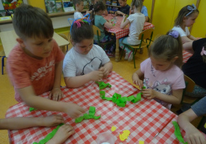 Dzieci siedzi przy stoliku i obijają patyczek zieloną bibułą