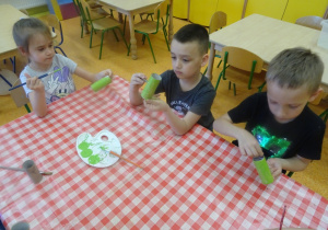 Troje dzieci siedzi przy stole i maluje zieloną farbą rolki od papieru toaletowego