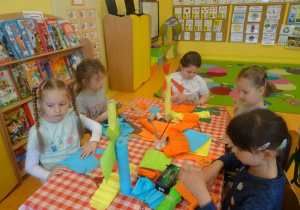 Pięcioro dzieci siedzi przy stole i wycinają, wyklejają elementy do makiety