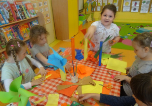 Pięć dziewczynek siedzi przy stoliku i wykonują wiatraki z rolek i kolorowego papieru