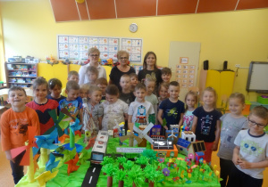 Grupa dzieci wraz z panią dyrektor i panią Arletą i Eweliną stoją za stolikiem , na którym jest makieta na temat "Moje ekologiczne miasto"wykonana przez dzieci