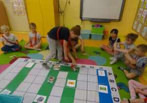Dzieci siedzą na dywanie mają na sobie szarfy w dwóch kolorach. Na środku leży mata do kodowania , Antek porusza się po wyznaczonym torze kubkiem w czerwonym kolorze