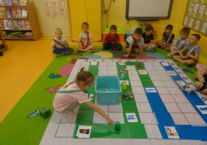 Dzieci siedzą na dywanie mają na sobie szarfy w dwóch kolorach. Na środku leży mata do kodowania , Kaja porusza się po wyznaczonym torze