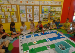 Dzieci siedzą na dywanie mają na sobie szarfy w dwóch kolorach. Na środku leży mata do kodowania , Kuba porusza się po wyznaczonym torze