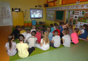 Dzieci siedzą na dywanie i oglądają film edukacyjny