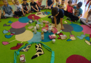 Dzieci siedzą na dywanie, jeden chłopiec siedzi w środku i wybiera produkty ekologiczne