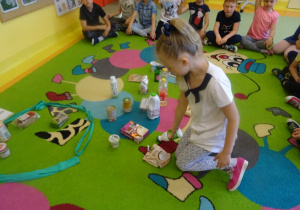 Dzieci siedzą na dywanie, jedna dziewczynka siedzi w środku i wybiera produkty ekologiczne