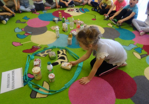 Dzieci siedzą na dywanie, jedna dziewczynka siedzi w środku i wybiera produkty ekologiczne, które wkłada do pętli