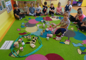 Dzieci siedzą na dywanie, jedna dziewczynka siedzi w środku i wybiera produkty ekologiczne