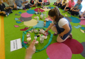 Dzieci siedzą na dywanie, jedna dziewczynka siedzi w środku i wybiera produkty ekologiczne, które wkłada do pętli