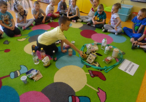 Dzieci siedzą na dywanie, jeden chłopiec siedzi w środku i wybiera produkty ekologiczne, które wkłada do pętli