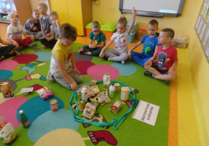 Dzieci siedzą na dywanie, jeden chłopiec siedzi w środku i wybiera produkty ekologiczne, które wkłada do pętli