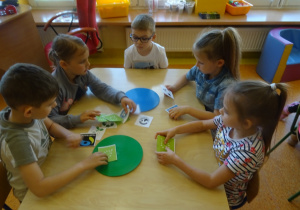 Pięcioro dzieci siedzi przy stoliku i wyszukują wśród wielu znaków ekologicznych
