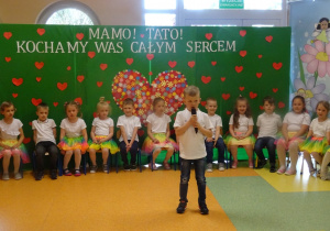 Dzieci siedzą w w półkolu, Marcel stoi na środku holu, trzyma mikrofon, mówi wiersz