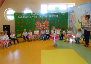Kalinka stoi na środku holu, trzyma mikrofon, mówi wiersz . Dzieci siedzą na krzesłach w półkolu obok stoi pani Dorota