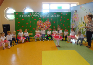 Maja stoi na środku holu, trzyma mikrofon, mówi wiersz . Dzieci siedzą na krzesłach w półkolu obok stoi pani Dorota