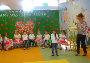 Igor stoi na środku holu, trzyma mikrofon, mówi wiersz . Dzieci siedzą na krzesłach w półkolu obok stoi pani Dorota