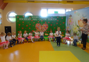 Marcel stoi na środku holu, trzyma mikrofon, mówi wiersz . Dzieci siedzą na krzesłach w półkolu obok stoi pani Dorota