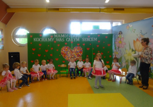 Malwina stoi na środku holu, trzyma mikrofon, mówi wiersz . Dzieci siedzą na krzesłach w półkolu obok stoi pani Dorota