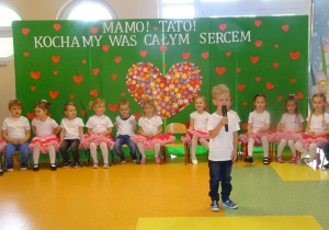 Chłopiec stoi na środku holu trzyma mikrofon, mówi wiersz, dzieci siedzą w półkolu na krzesłach