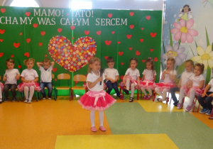 Dziewczynka stoi na środku holu trzyma mikrofon, mówi wiersz, dzieci siedzą w półkolu na krzesłach