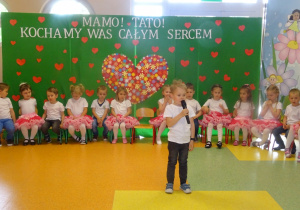 Chłopiec stoi na środku holu trzyma mikrofon, mówi wiersz, dzieci siedzą w półkolu na krzesłach