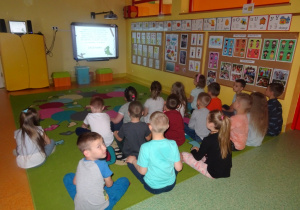 Dzieci siedzą na dywanie i oglądają na tablicy interaktywnej film o szkodliwości palenia papierosów