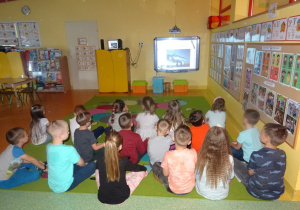 Dzieci siedzą na dywanie i oglądają na tablicy interaktywnej film jak fabryki zanieczyszczją powietrze