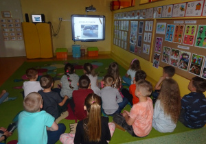Dzieci siedzą na dywanie i oglądają na tablicy interaktywnej film co dymi