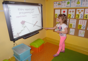 Julka stoi przed tablicą interaktywną i pokazuje wskaźnikiem na tablicy róże