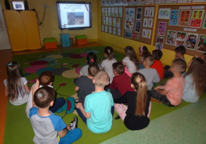Dzieci siedzą na dywanie i oglądają na tablicy interaktywnej film gdzie nie wolno palić papierosów