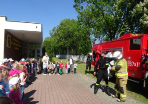 Dzieci stoją w półkolu przed nimi stoi wóz strażacki i dwóch strażaków