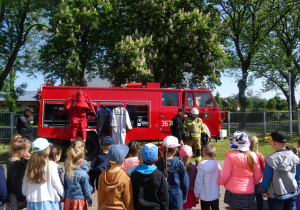 Dzieci stoją w półkolu przed nimi stoi wóz strażacki