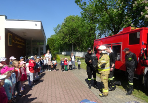 Dzieci stoją w półkolu przed nimi stoi wóz strażacki i czworo strażaków