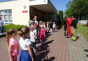 Dzieci stoją w półkolu z paniami, przed nimi stoją strażacy, jeden z nich pokazuje strój strażaka
