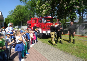 Dzieci stoją i oglądają jak strażacy puszczją wodę z węża strażackiego
