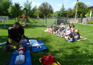 Dzieci siedzą na kocach, strażacy pokazują na fantomach jak udzielać pierwszej pomocy przedmedycznej