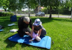 Dziewczynka pokazuje jak udzielić pierwszej pomocy małemu dziecku, obok przyklęka strażak