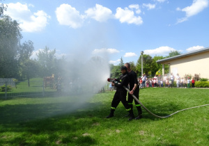Dwóch strażaków pokazuje jak z węża strażackiego leci woda, dzieci stoją w oddali