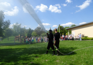 Dwóch strażaków pokazuje jak z węża strażackiego leci woda, dzieci stoją w oddali