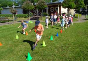 Dzieci stoją ustawieni w dwa rzędy pierwsi z rzędów biegną slalomem między pachołkami do chorągiewek