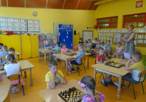Sześć par gra w szachy, pani Arletka stoi pozostałe dzieci siedzą na krzesłach