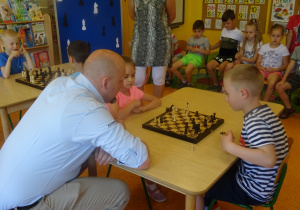 Kaja i Gabryś grają w szachy przy ich stoliku przykucnął pan