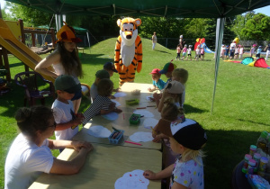 Dzieci stoją wokół stolików rysują na chmurce, przygląda się im Tygrysek