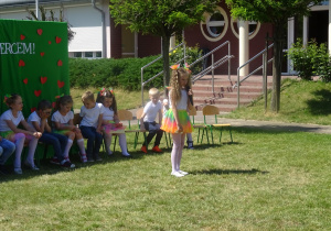 Magda stoi na środku, trzyma mikrofon, mówi wiersz. Dzieci siedzą na krzesłach
