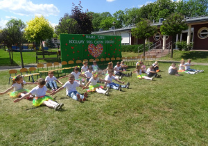 Dzieci siedzą w siadzie prostym na trawie trzymają ręce w bok